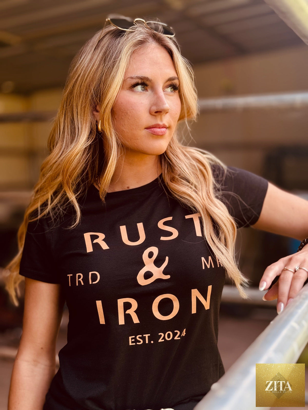 Alt text: Woman wearing a black "High Noon" T-shirt by Rust & Iron Clothing, with blonde hair and sunglasses on her head, outdoors.